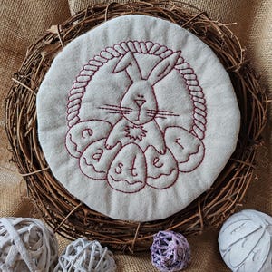 May include: A white fabric circle with red embroidery of a bunny rabbit holding Easter eggs that spell out "Easter". The circle is surrounded by a brown twig wreath.