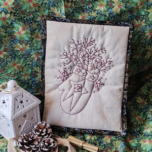 May include: A cream-coloured, quilted Christmas mitten with an embroidered design of candy canes, snowflakes, and a gingerbread man. The mitten is framed with a black border and set against a festive floral background. A white ceramic lantern and pine cones are in the foreground.