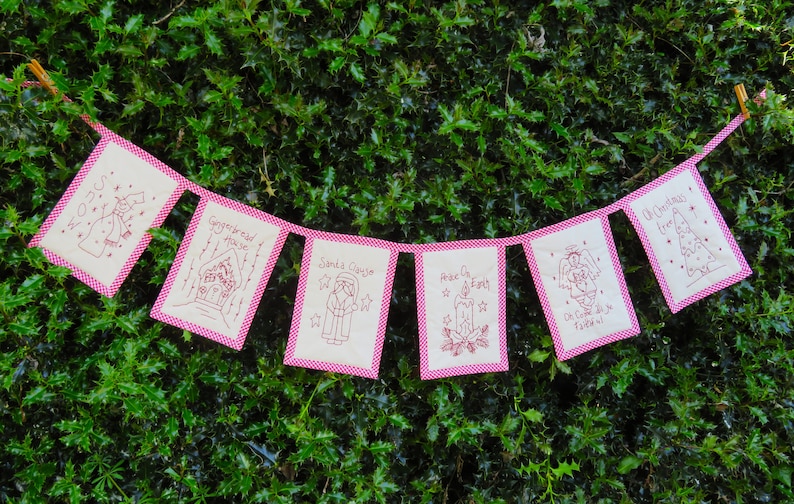 May include: A string of six festive fabric pennants with pink borders and red embroidery. The pennants feature Christmas-themed designs, including a gingerbread house, Santa Claus, a Christmas tree, and an angel. The text on the pennants reads "Gingerbread House", "Santa Clause", "Peace on Earth", "Oh Christmas Tree", and "An Angel of the Lord".