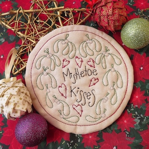 May include: A round, cream-coloured fabric ornament with green embroidered mistletoe sprigs and red embroidered hearts. The text "Mistletoe Kisses" is embroidered in black thread.