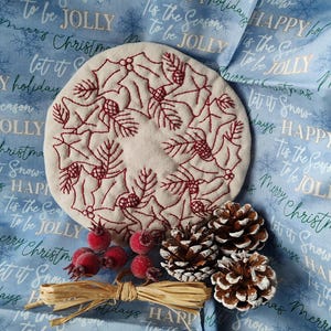 May include: A white fabric circle with red embroidery of holly leaves, berries, and pine cones. The fabric circle is on a blue background with the text "Happy Holidays" and "Tis the Season to be Jolly" repeated. There are also pine cones and red berries on the fabric.