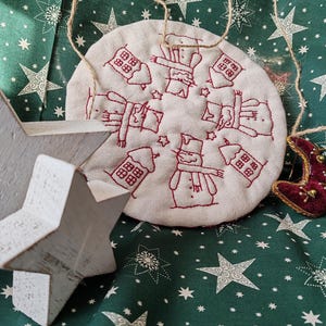 May include: A white fabric circle with red embroidery of snowmen, houses, and stars. The fabric is on a green background with white stars.
