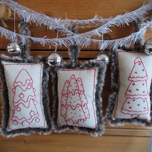May include: Three handmade Christmas tree decorations with a white background and red embroidery. Each decoration is trimmed with brown faux fur.