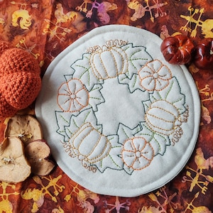 Op de afbeelding: Een ronde, gebroken witte stoffen mat met een geborduurde krans van oranje pompoenen en groene bladeren. Het borduurwerk maakt gebruik van eenvoudige rijgsteken om de vormen van de pompoenen en het gebladerte in een cirkelvormig ontwerp te omlijnen.