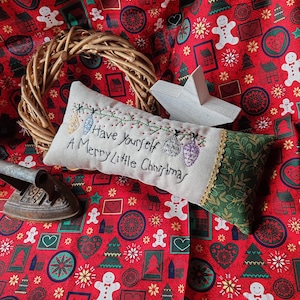 Könnte beinhalten: Ein weißes Kissen mit grünem Besatz und dem aufgestickten Text "Have Yourself A Merry Little Christmas". Das Kissen liegt auf einem roten Stoff mit einem sich wiederholenden Muster aus Weihnachtsbäumen, Lebkuchenmännern, Schneeflocken und Herzen.