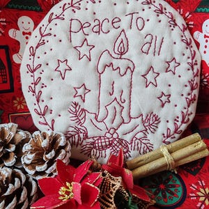 May include: A circular, cream-coloured Christmas decoration embroidered with the words "Peace To All" and a candle design in red thread. The decoration is surrounded by a red floral pattern, pine cones, and cinnamon sticks.