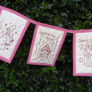 May include: A red and white gingham fabric banner with three embroidered panels. The panels feature a snowman, a gingerbread house, and Santa Claus. The banner is hanging on a green bush.
