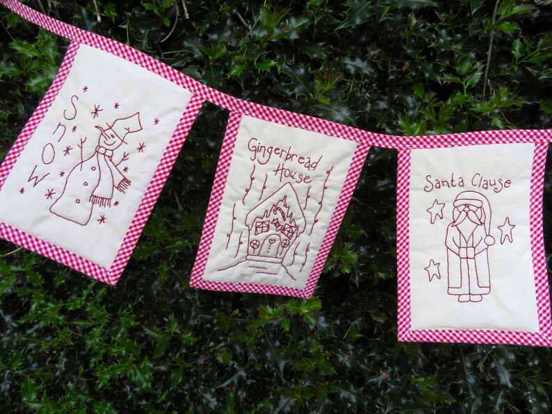 May include: A red and white gingham fabric banner with three embroidered panels. The panels feature a snowman, a gingerbread house, and Santa Claus. The banner is hanging on a green bush.