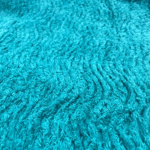 Könnte beinhalten: Nahaufnahme eines strukturierten, türkisfarbenen Stoffes. Das Material hat ein weiches, plüschiges Aussehen mit einer welligen, unebenen Oberfläche. Die Farbe ist ein lebendiges, gesättigtes Türkis, das eine visuell ansprechende Textur erzeugt.