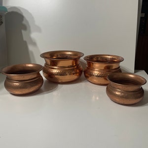 May include: Four copper-coloured pots of varying sizes are arranged on a white surface. The pots have a rounded shape with a flared rim and a hammered texture. The pots are empty and appear to be decorative.