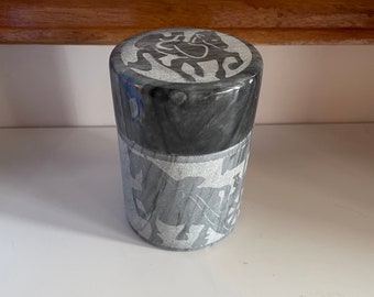 May include: A gray stone cylindrical container with a carved design of a rider on a horse on the lid.