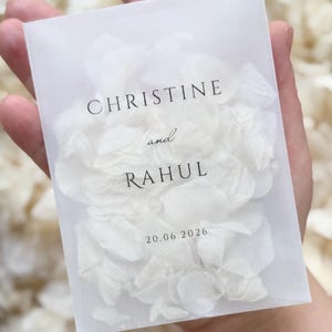 Könnte beinhalten: Ein rechteckiger, halbtransparenter Umschlag aus Pergamentpapier, gefüllt mit weißen Blütenblättern. Auf der Vorderseite ist der schwarze Text "CHRISTINE and RAHUL" sowie das Datum "20.06.2026" in einer klaren Serifenschrift zentriert aufgedruckt.