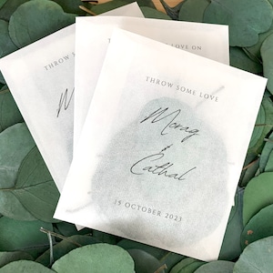 Op de afbeelding: Witte papieren confetti met de tekst "Throw Some Love" en de namen "Morag & Cathal" in cursief schrift. De datum "15 oktober 2023" staat onder de namen gedrukt.