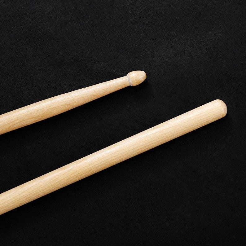 May include: Two wooden drumsticks with rounded tips, lying diagonally against a black background. The drumsticks are light brown and appear to be made of wood. The tips are slightly darker and rounded.