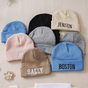 Peut inclure: Plusieurs bonnets personnalisés de différentes couleurs, dont bleu, rose, beige et noir. Chaque bonnet porte un nom brodé en fil blanc. Les noms incluent "BOSTON", "ALLIE", "SALLY" et "JENSEN".