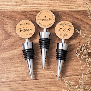 May include: Three wooden bottle stoppers with personalized engraved text. The stoppers are silver with black rubber tops. The text on the stoppers reads "Mr. & Mrs. Fishers June 17th, 2025", "Steve & Rachel married October 15, 2024", and "L & G".