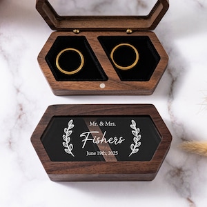 Op de afbeelding: Een houten ringendoos met een doorzichtig deksel. De doos heeft twee vakken voor ringen en een zwart plaatje met de tekst "Mr. & Mrs. Fishers June 19th, 2025" en een laurierkransontwerp.