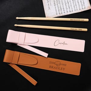 May include: Two pairs of wooden drumsticks with text, "Best drummer ever" and "I love you - Sally". Two personalized drumstick cases, one pink with "Cerelia" and one brown with "BRADLEY" and a drumstick graphic.
