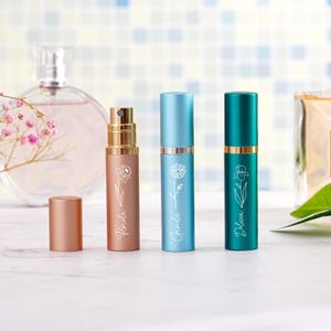 May include: Three cylindrical metal perfume atomizers in rose gold, light blue, and dark teal. Each features a white line-art floral illustration and a name: Florida, Camila, and Delicia. A matching rose gold cap sits beside the first atomizer.