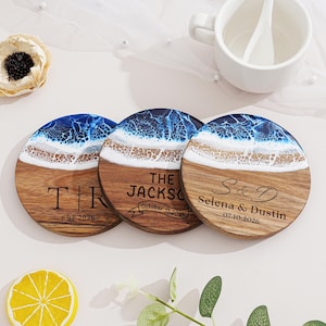 Könnte beinhalten: Drei runde Untersetzer aus Holz mit Strandmotiv. Die obere Hälfte zeigt ein blau-weißes Harzwellenmuster, während die untere Hälfte aus Naturholz besteht. Jeder Untersetzer hat personalisierten Text, einschließlich Initialen und Daten.