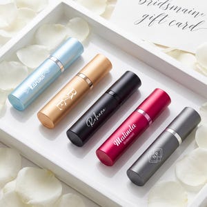 May include: Five cylindrical perfume atomizers in light blue, gold, black, magenta, and dark gray, each featuring custom white text or monograms. The atomizers are arranged diagonally in a white tray, accompanied by a card labeled Bridesmaid gift card.