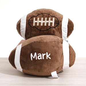 May include: Two brown plush footballs with white stripes and embroidered details. One football has the name "Mark" in white. The other has an embroidered design resembling football laces. These plush footballs are perfect for sports fans.