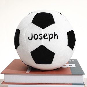 Può includere: Un pallone da calcio di peluche bianco e nero, con il nome "Joseph" stampato in nero. La palla è appoggiata su una pila di libri. Il pallone da calcio ha un design pentagonale classico.