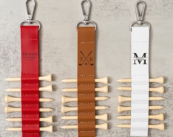 May include: Three personalized golf tee holders with leather straps and metal clips. The holder on the left is red and says "Howard  Groomman 12.20.2023". The middle holder is brown and has a golf club and ball design. The holder on the right is white and says "Matthew M".