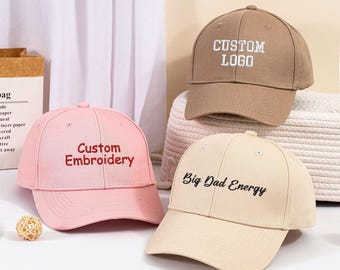 May include: Three baseball caps with different embroidered text. The first cap is brown with white text that says "CUSTOM LOGO". The second cap is pink with red text that says "Custom Embroidery". The third cap is beige with black text that says "Big Dad Energy".