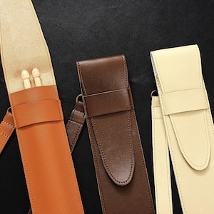 May include: Three leather drumstick cases in shades of tan, brown, and cream. The tan case is open, revealing two wooden drumsticks. Each case has a strap and a buckle closure. The background is black.