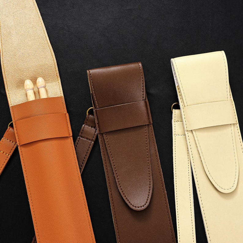 May include: Three leather drumstick cases in shades of tan, brown, and cream. The tan case is open, revealing two wooden drumsticks. Each case has a strap and a buckle closure. The background is black.