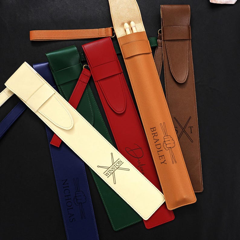 May include: A collection of personalized leather drumstick cases in various colors, including cream, red, green, brown, and tan. Each case features custom text and drumstick graphics. Some cases are open, revealing wooden drumsticks.