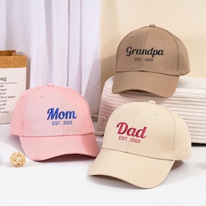 Peut inclure: Trois casquettes de baseball avec du texte brodé. Les casquettes sont marron, rose et beige. Le texte sur les casquettes est "Grandpa EST. 2025", "Mom EST. 2025" et "Dad EST. 2025".