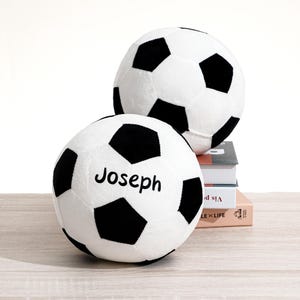 May include: Two plush soccer balls, white with black pentagons, one with the name "Joseph" printed on it. One ball rests on a stack of books, the other is in the background. The balls are on a light wood surface.