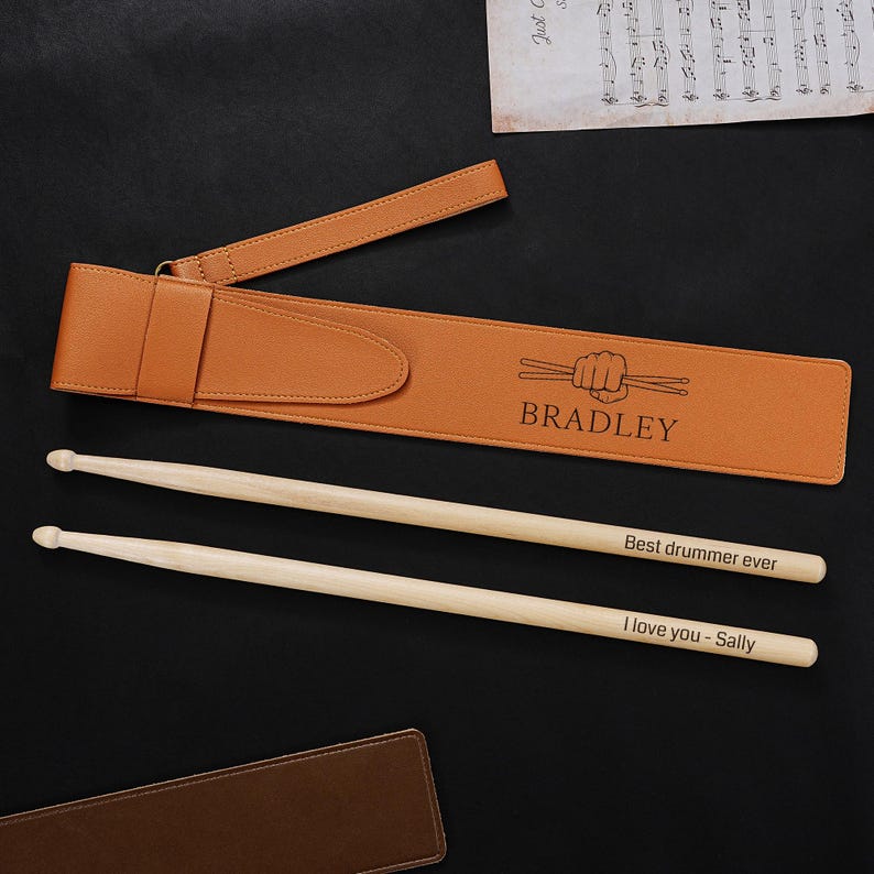 May include: A tan leather drumstick case with the name "Bradley" and a drumstick graphic. Two wooden drumsticks are next to the case, one with "Best drummer ever" and the other with "I love you - Sally" engraved on them. The background is black.
