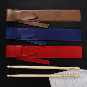 May include: Three leather drumstick cases in brown, blue, and red, each with a strap and personalized text. Two wooden drumsticks are visible, one with "Mickey" and the other with "No Drums, No Life" printed on them. Sheet music is in the foreground.
