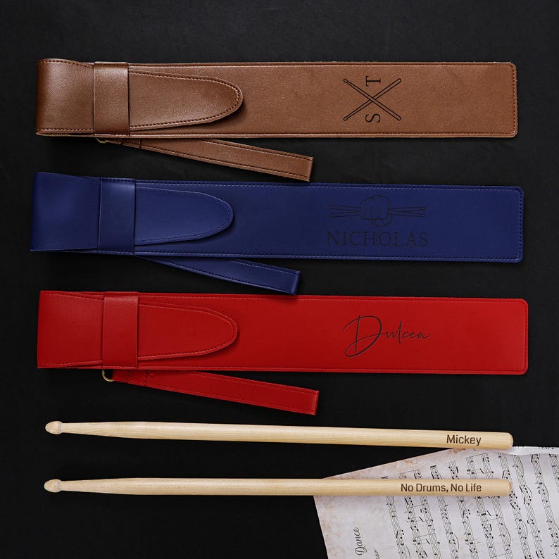 May include: Three leather drumstick cases in brown, blue, and red, each with a strap and personalized text. Two wooden drumsticks are visible, one with "Mickey" and the other with "No Drums, No Life" printed on them. Sheet music is in the foreground.