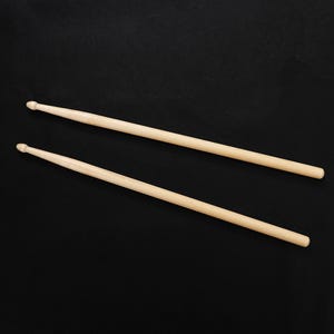 May include: Two wooden drumsticks, light brown in color, are positioned diagonally against a black background. Each stick has a rounded tip on one end and tapers to a thinner handle. The sticks are parallel to each other.