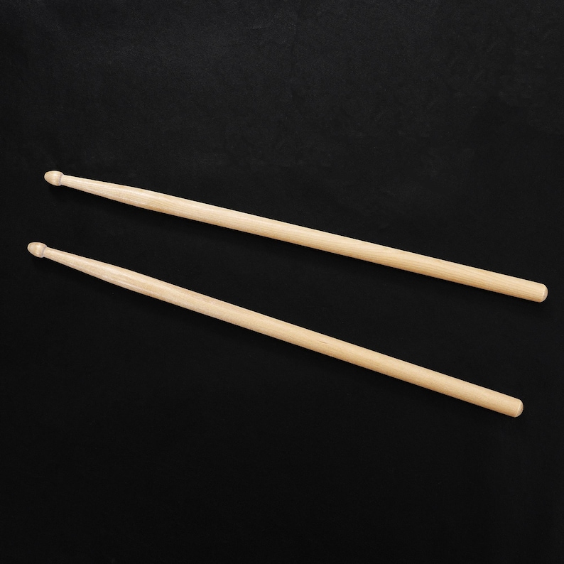 May include: Two wooden drumsticks, light brown in color, are positioned diagonally against a black background. Each stick has a rounded tip on one end and tapers to a thinner handle. The sticks are parallel to each other.