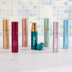 Könnte beinhalten: Sieben zylindrische Parfümzerstäuber aus Metall in verschiedenen Farben wie Gold, Magenta, Roségold, Petrol, Minzgrün, Hellrosa und Hellblau. Jede Flasche ist mit individuellem weißem Text oder floralen Grafiken und einem Sprühkopf versehen.