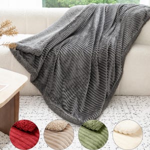 May include: A grey, plush blanket with a ribbed design is draped over a white sofa. The blanket has a soft, fuzzy texture and a contrasting border. Four colour swatches at the bottom show red, beige, green, and white blanket options.