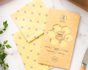 May include: Beeswax food wraps in yellow with purple flower patterns, alongside fresh vegetables like carrots, red cabbage, and red onion. A knife and a package with text are also visible.