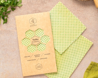 May include: Beeswax food wraps with green polka dots are displayed next to a package with the text "Vaško drobelės". The wraps are on a light brown surface with ingredients like pumpkin seeds, lime slices, and a butternut squash half.