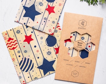 May include: Beeswax wraps with a festive design featuring red stars, blue ornaments, and snowflakes. The wraps are displayed with a brown package that reads "Bees wax wraps" and "Assorted 3 pack". The image also includes Christmas ornaments and greenery.