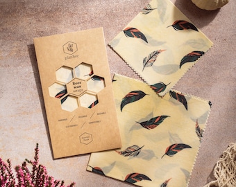 May include: Beeswax food wraps with a feather pattern in a kraft paper package. The package reads "planBee Bees wax wraps" and includes the words "handmade, reusable, washable, eco-friendly, compostable". Two small white pumpkins and a pink flowering plant are also in the image.