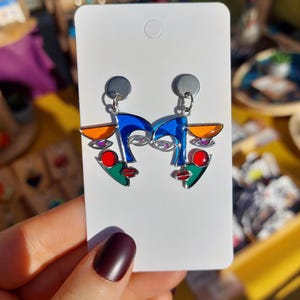 May include: A pair of silver earrings with an abstract face design in blue, green, red and orange.