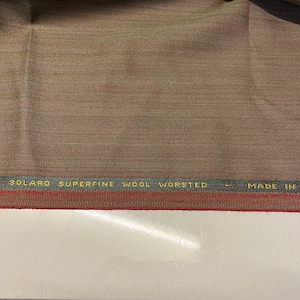 Op de afbeelding: Close-up van een bruine stof met een rode en groene streep. De stof heeft de tekst "ORIGINAL SOLARO SUPERFINE WOOLWORTED - MADE IN ENGLAND" erop gedrukt.