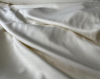 May include: Close-up of off-white fabric with visible texture and folds. The fabric appears to be a woven material, possibly linen or cotton, with a subtle sheen. The folds create shadows and highlights.