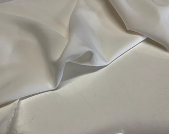 May include: Close-up of ivory-colored fabric with soft folds and subtle shadows. The fabric appears smooth and possibly silky, with a slight sheen. The material is draped, creating a sense of texture and depth. Ideal for apparel or home decor.