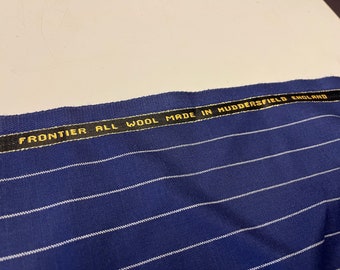 May include: Close-up of a blue fabric with white stripes. The fabric has a black label with gold lettering that reads "FRONTIER ALL WOOL MADE IN HUDDERSFIELD ENGLAND".